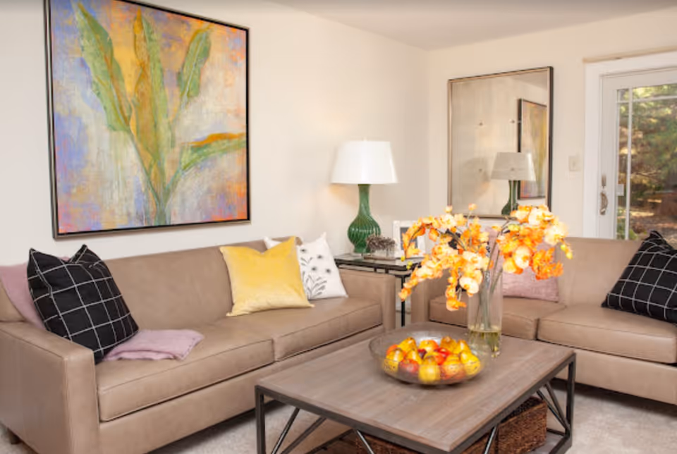 Stylish living room with two beige sofas, a wooden coffee table holding a bowl of fruit and a vase of orange flowers, decorative pillows, lamps, and wall art.