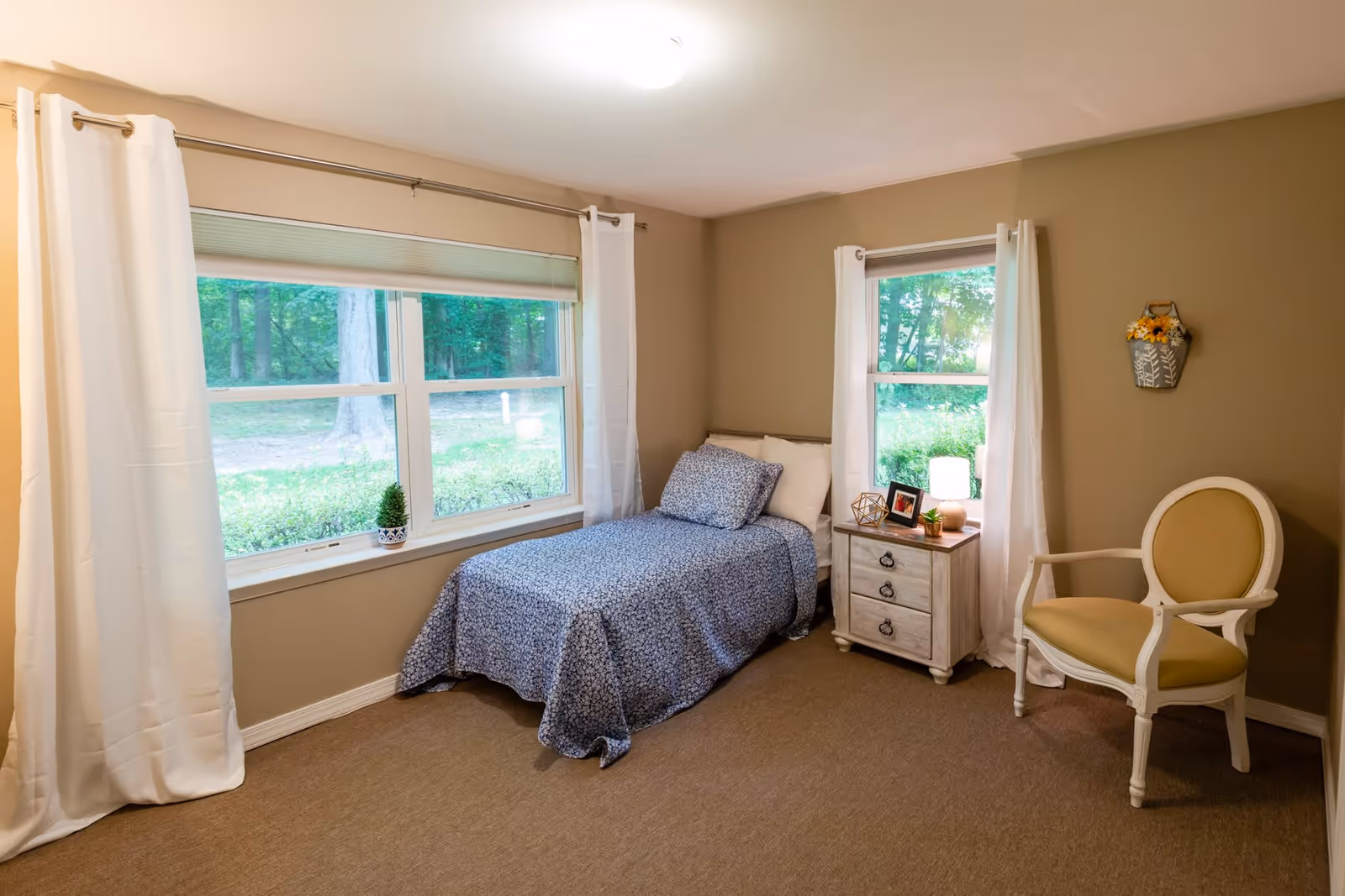 A cozy bedroom with a single bed covered in a blue patterned bedspread and matching pillow. The room has beige walls and carpet, two windows with white curtains, a small wooden nightstand with a lamp, framed photo, and decorative items. There is also a beige upholstered armchair and a small wall decoration with sunflowers.