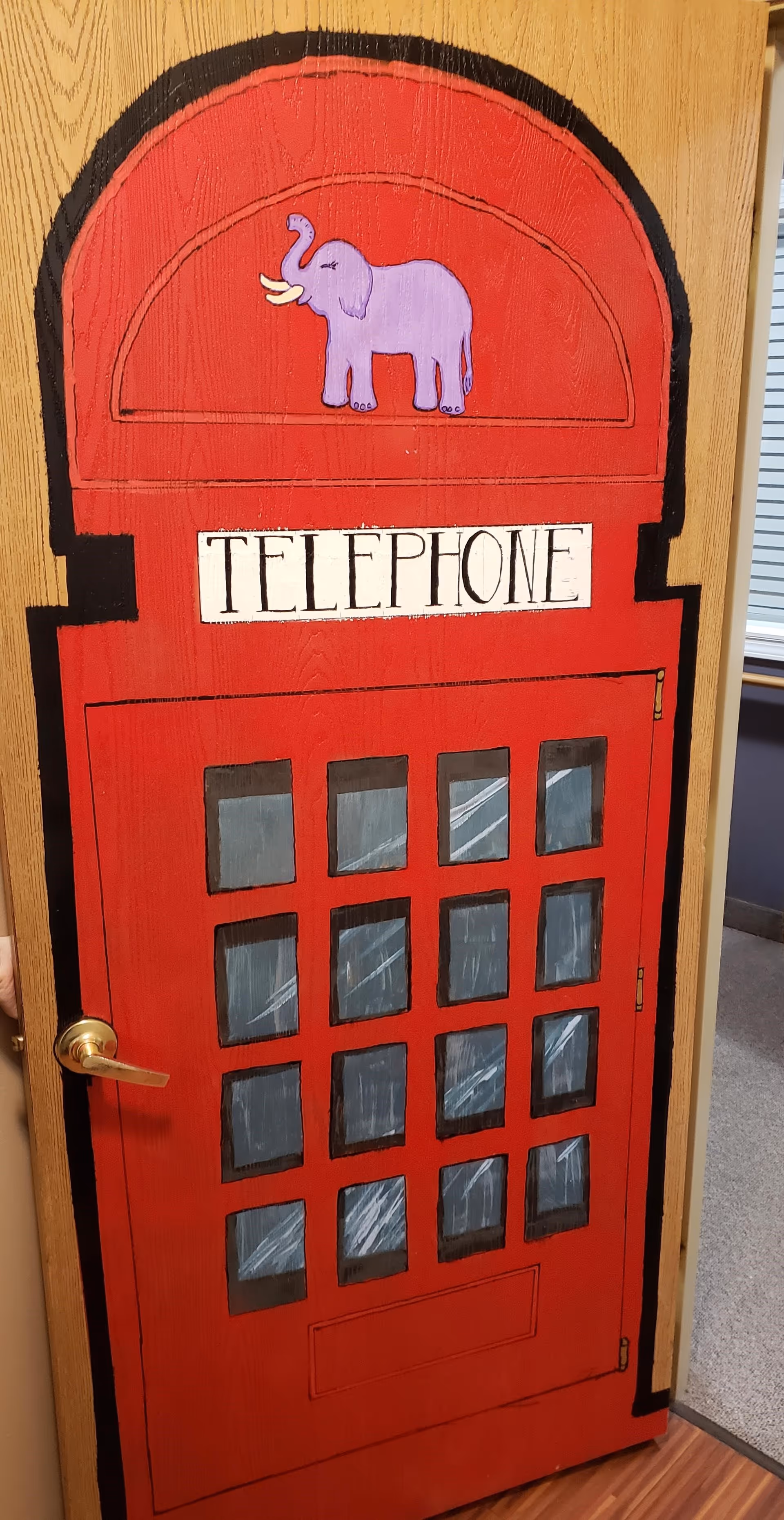 A wooden door painted to resemble a red British telephone booth with a purple elephant illustration above the word TELEPHONE. The door has a gold handle and is partially open, showing a glimpse of a carpeted room beyond.