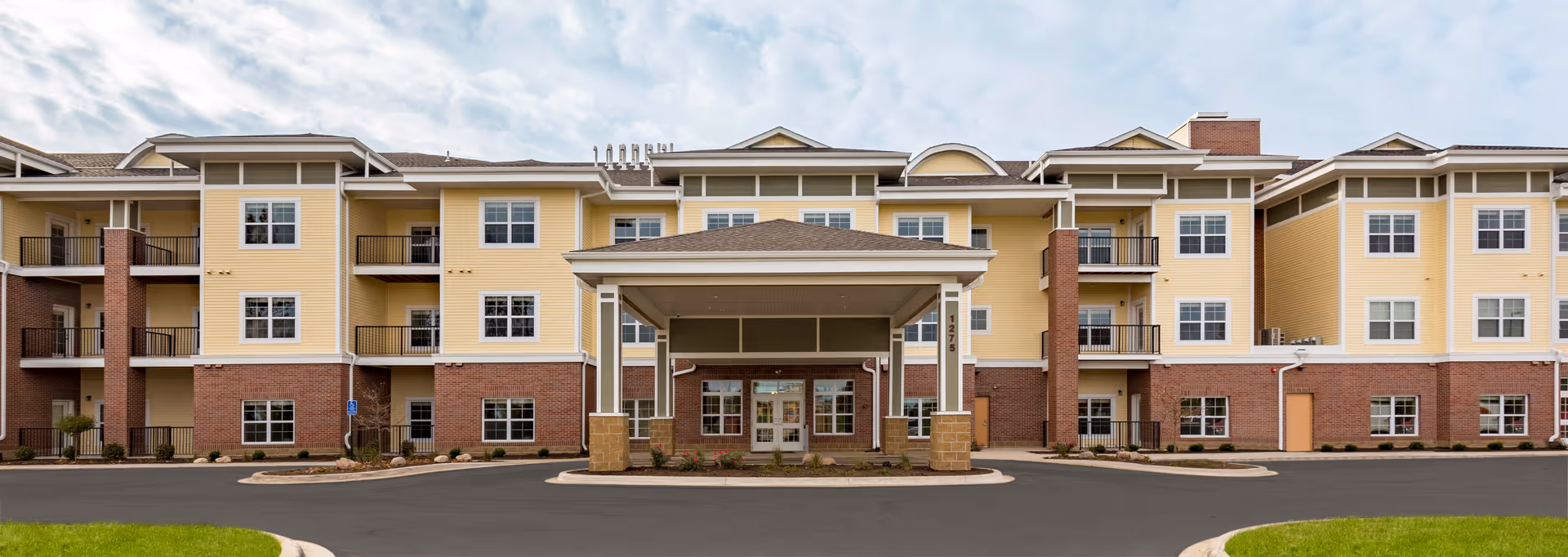 Front exterior view of a three-story senior living facility building with yellow siding and red brick accents. The building features multiple windows and balconies, with a covered entrance in the center. The driveway and landscaped areas are visible in the foreground under a partly cloudy sky.