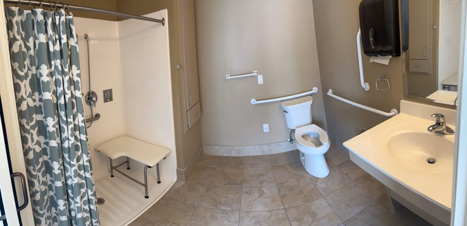 A spacious bathroom with beige walls and tiled floor, featuring a walk-in shower with a patterned blue and white curtain and a foldable shower seat, a white toilet with grab bars on the walls around it, a white sink with a chrome faucet, a large mirror, and a black paper towel dispenser mounted on the wall.