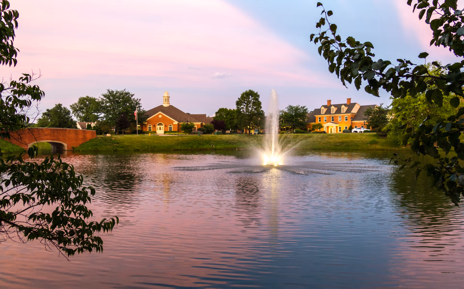A lit fountain in a pond at dusk with brick buildings, a small bridge, and trees along the shoreline.