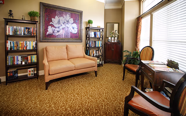A cozy sitting area in a senior living facility with a beige loveseat centered between two black bookshelves filled with books. Above the loveseat hangs a large framed painting of white flowers on a purple background. To the right, there is a wooden desk with two chairs, a plant, and large windows with white blinds letting in natural light. The room has patterned carpet and beige walls.