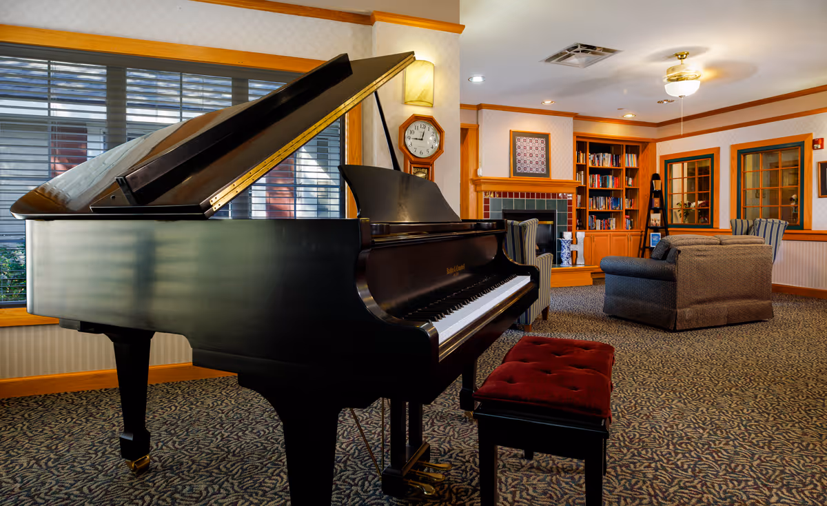 Interior of a cozy living room in an assisted living facility featuring a black grand piano with a red cushioned bench, a fireplace with a tiled surround, built-in wooden bookshelves filled with books, comfortable upholstered chairs and sofas, patterned carpet, and windows with blinds.