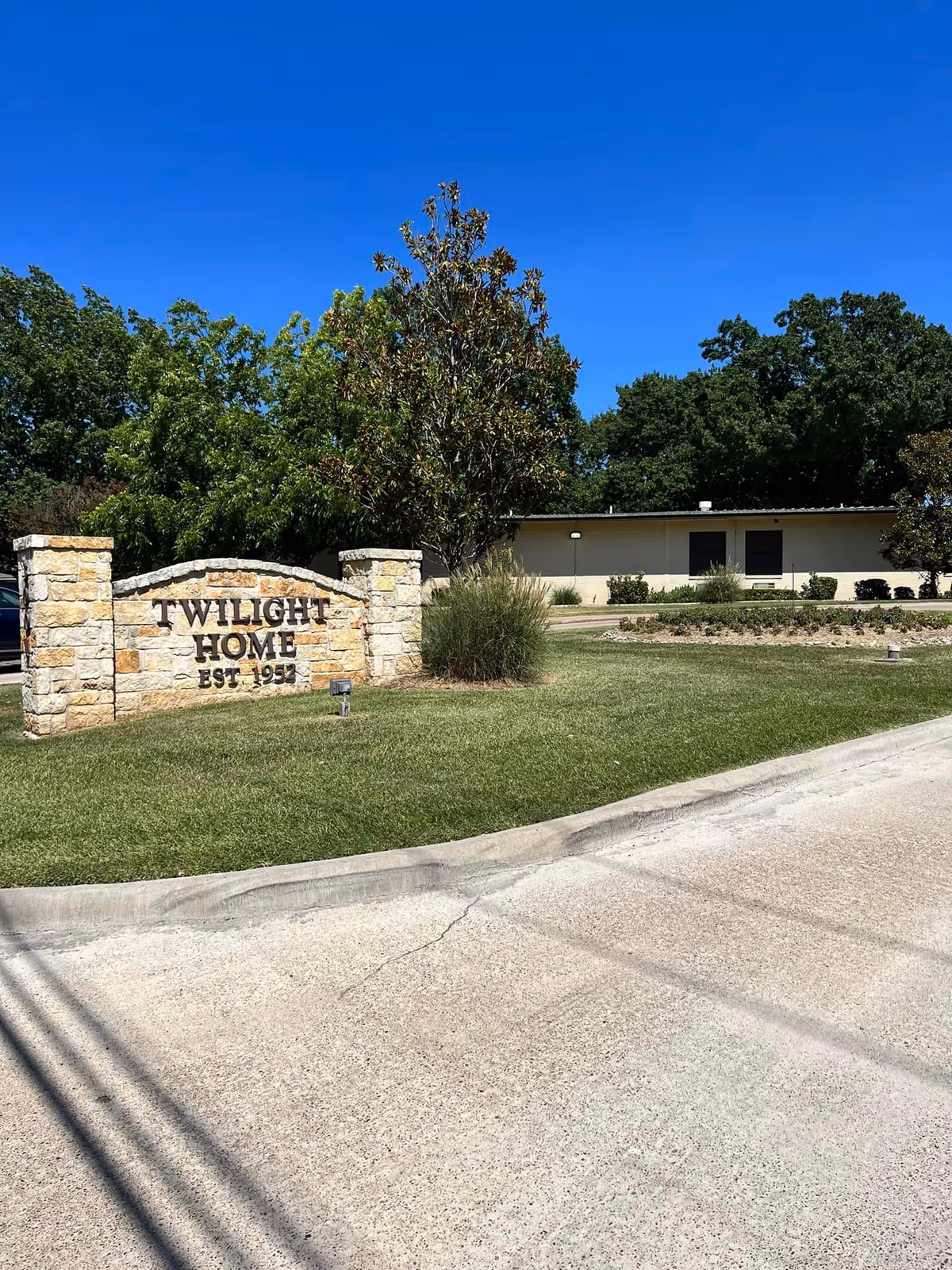 A stone sign reading "Twilight Home EST. 1951" on a grassy lawn in front of a low building with trees and a clear blue sky.