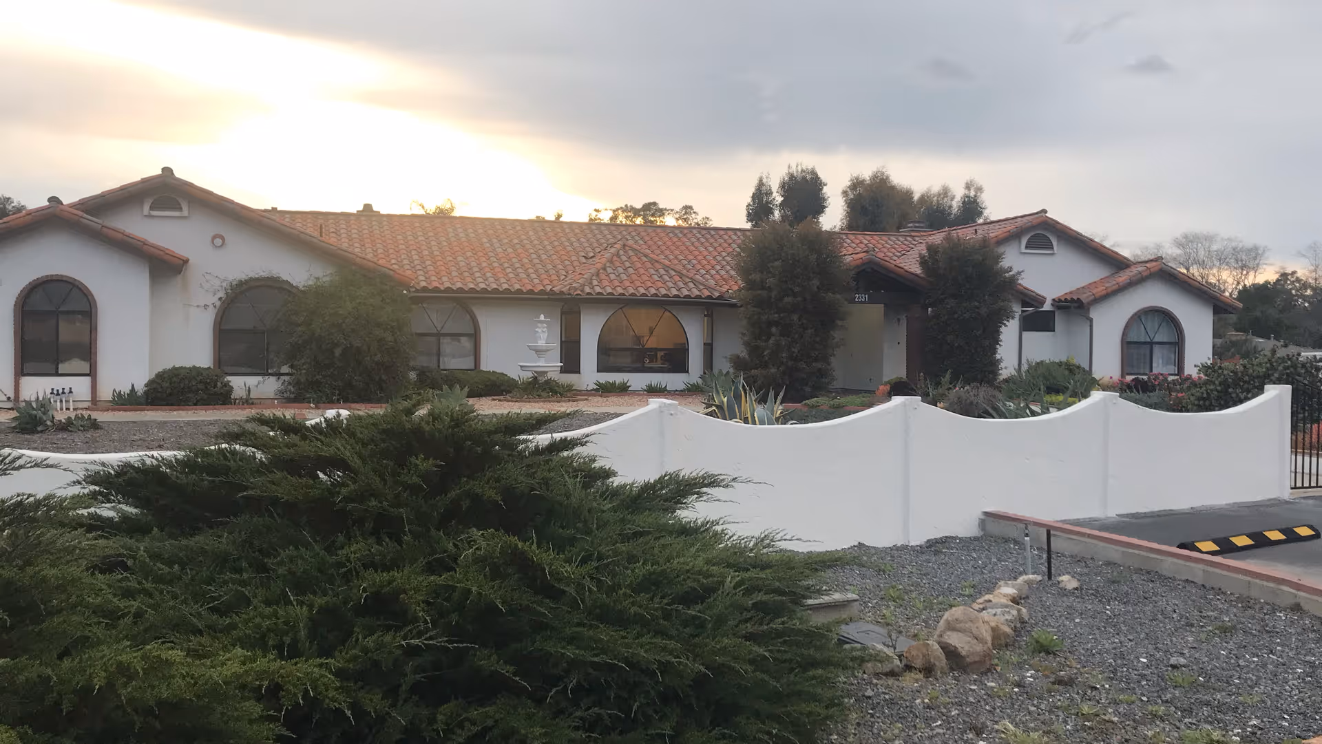 Exterior view of Monte Vista Manor, a single-story building with white walls and a red tile roof, surrounded by greenery and a white curved wall in front. The sky is cloudy with the sun setting in the background.