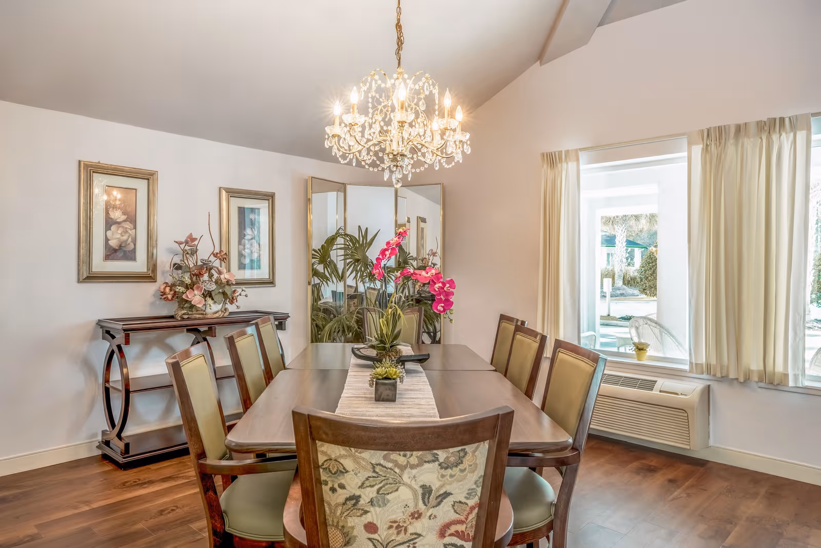A well-lit dining room with a wooden dining table surrounded by eight chairs, a chandelier hanging above, floral decorations on the table and a side table, two framed floral artworks on the wall, a large mirror, and a window with cream-colored curtains letting in natural light.