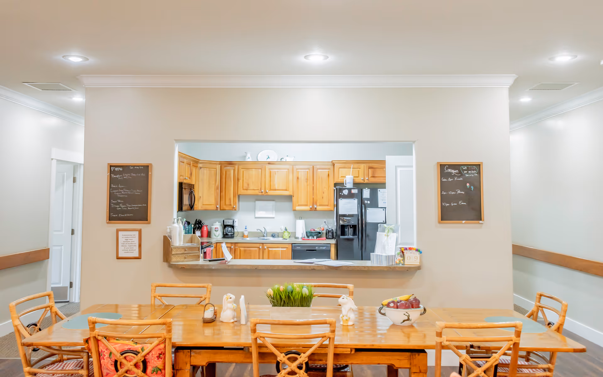A dining area with a wooden table and six chairs in front of a kitchen pass-through window. The kitchen has wooden cabinets, a black refrigerator, a microwave, and various kitchen appliances. Two chalkboards with handwritten notes are mounted on the wall on either side of the pass-through window. The table is decorated with small bunny figurines, a bowl of fruit, and a small plant.
