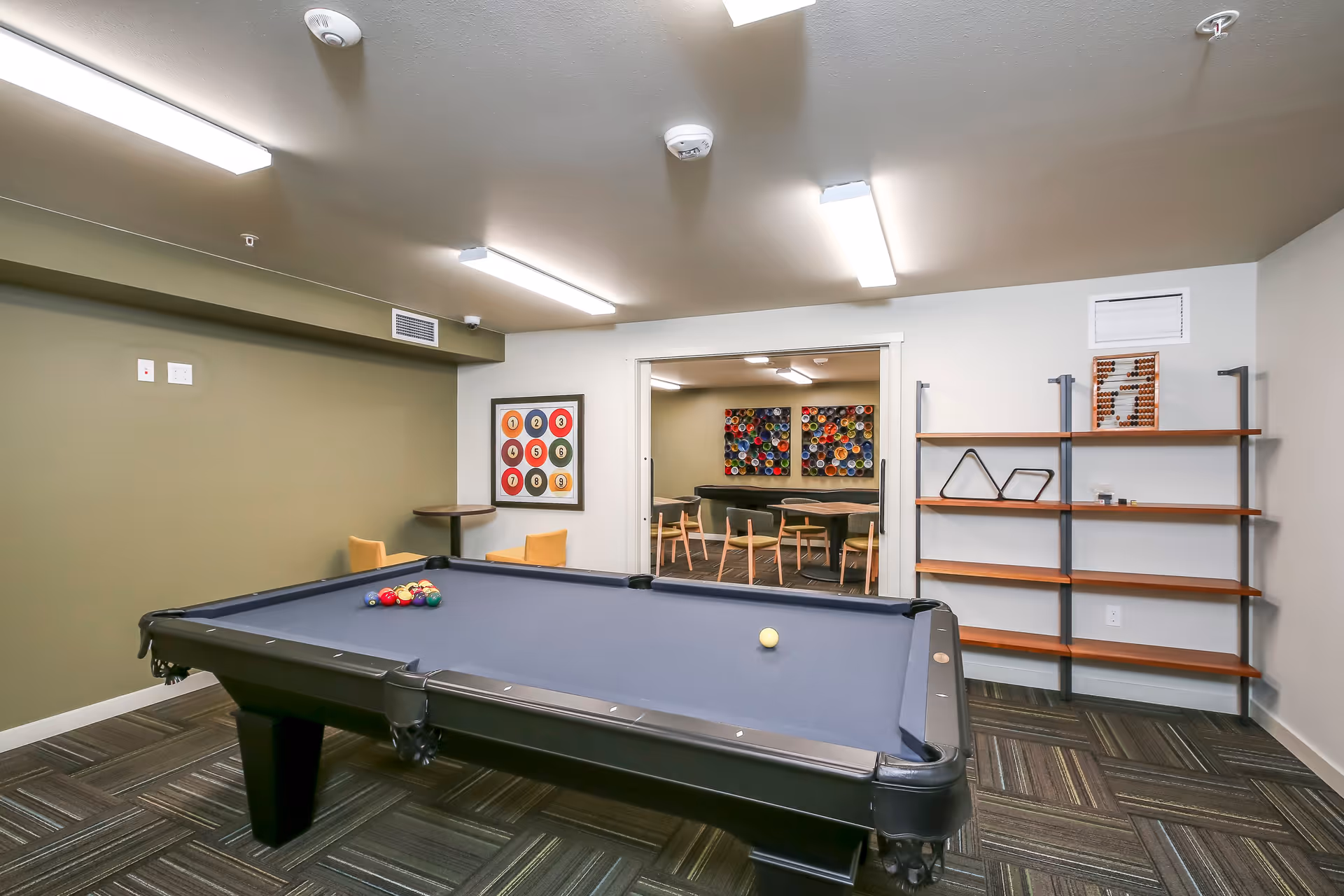 Interior room with a pool table in the foreground, a green accent wall, and a shelving unit on the right. In the background, there is an adjoining room with tables and chairs, colorful wall art, and bright overhead lighting.