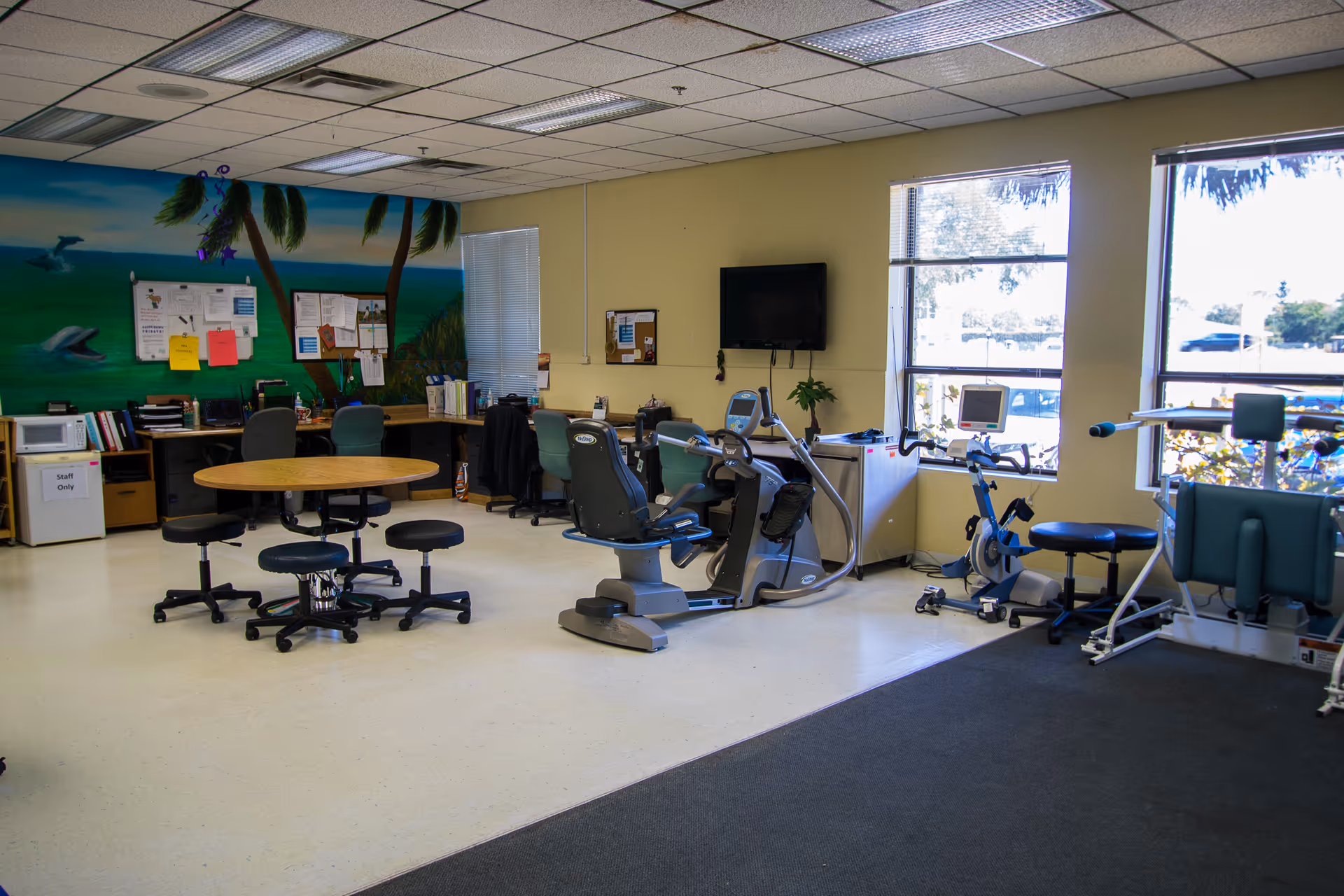 A rehabilitation or therapy room with exercise equipment including stationary bikes and other fitness machines. There is a round table with stools in the center, desks with chairs and office supplies along the back wall, and large windows letting in natural light. The walls are painted yellow and one wall features a mural with palm trees and ocean scenery.