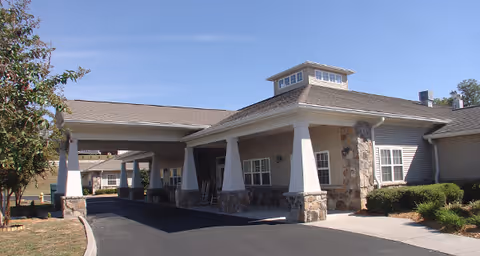 Exterior view of a single-story senior living facility building with a covered entrance supported by large white columns with stone bases. The building has beige siding, multiple windows, and a small cupola on the roof. There is a paved driveway leading up to the entrance, surrounded by some landscaping with bushes and a tree.