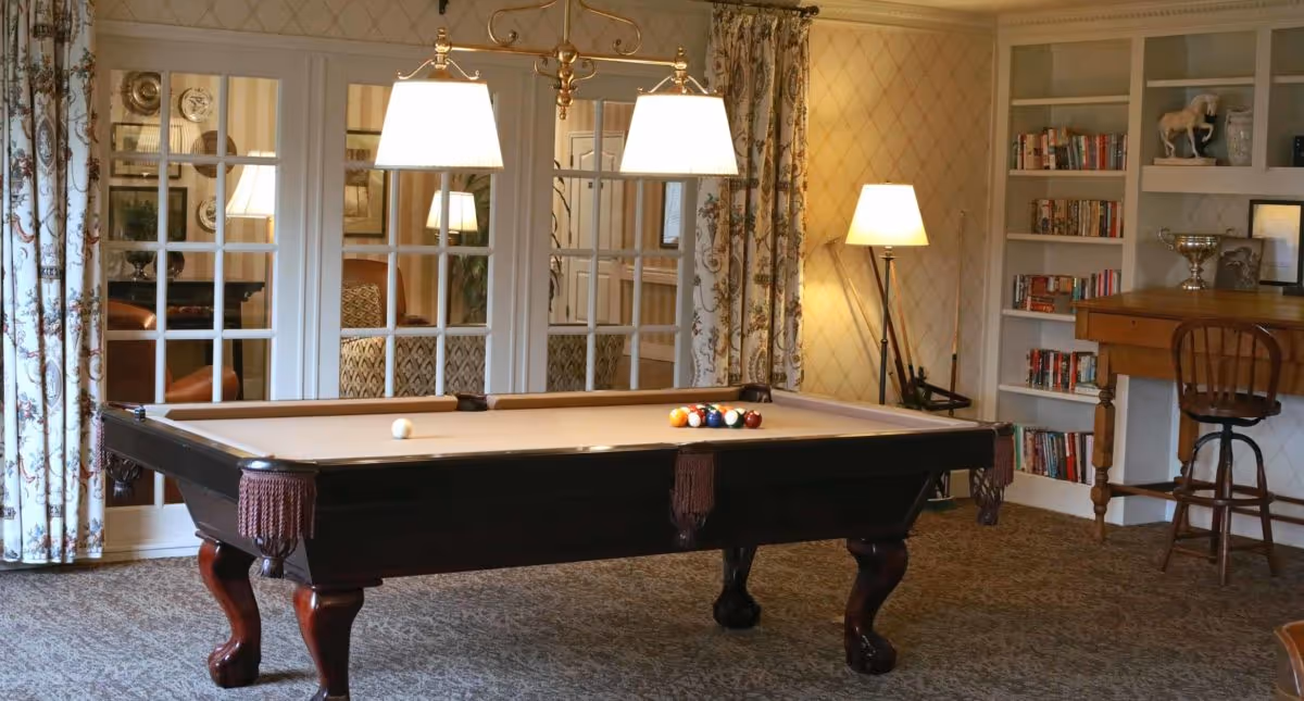 A cozy interior room featuring a billiards table with balls set up for a game. The room has patterned curtains, a carpeted floor, a floor lamp, and built-in bookshelves filled with books and decorative items. There is also a wooden desk with a chair.