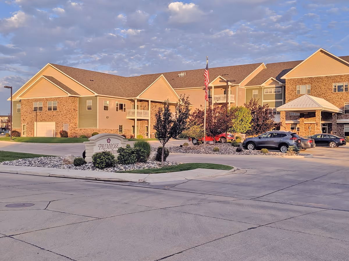 Exterior view of Primrose Retirement Community of Gillette showing a large, multi-story building with beige and green siding and brick accents. There are several cars parked in front, a landscaped area with small trees and bushes, and an American flag on a flagpole. The sky is partly cloudy with soft sunlight illuminating the scene.