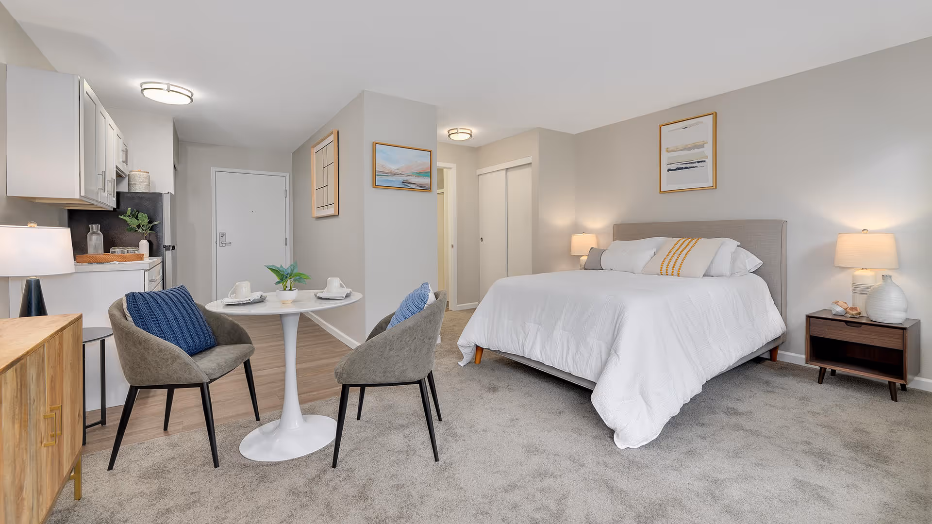 Studio apartment bedroom with a bed, two nightstands, a small round dining table with two chairs, and a kitchenette.