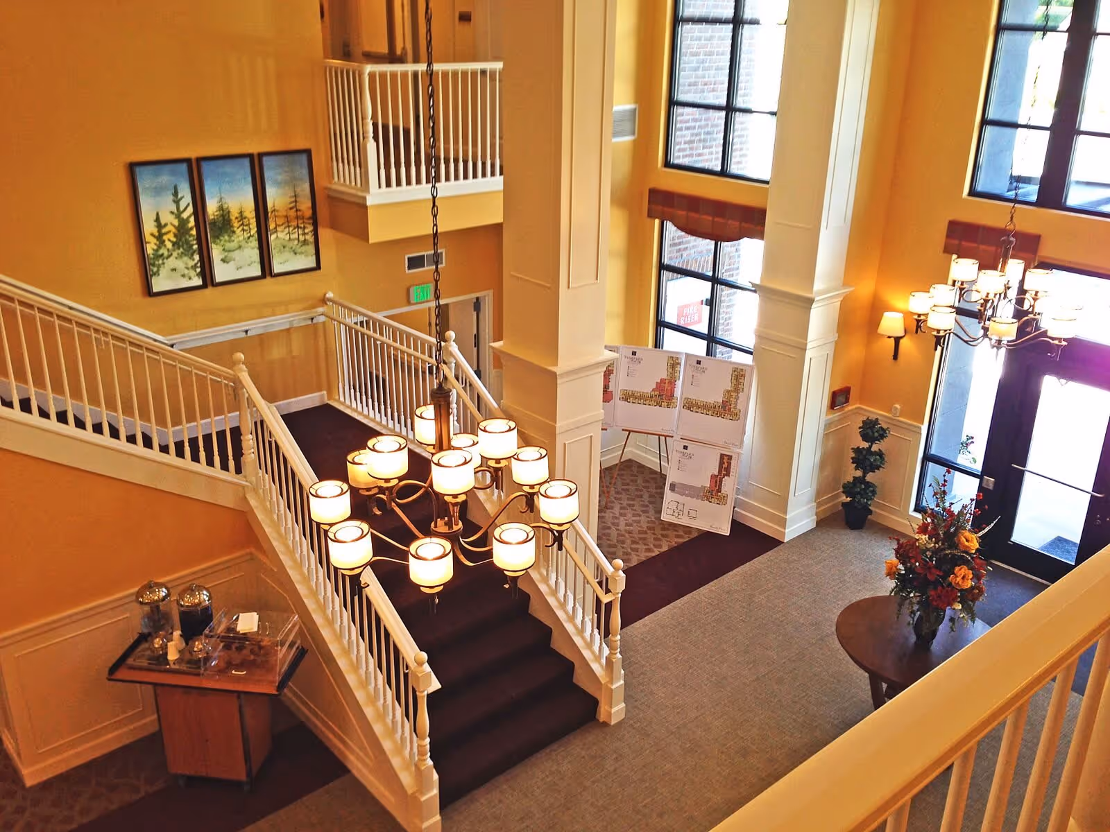 Interior view of a senior living facility lobby with a staircase featuring white railings and dark carpet. A large chandelier with multiple lights hangs above the stairs. There are large windows letting in natural light, a round table with a floral arrangement, and floor plans displayed on easels near the entrance. The walls are painted a warm yellow color, and there is a small table with refreshments near the staircase.