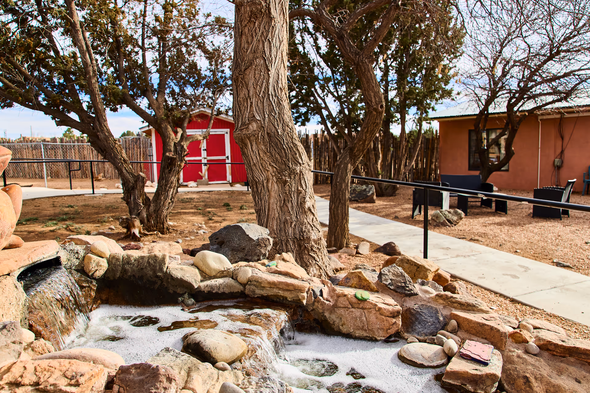 Outdoor courtyard with a small rocky water feature in the foreground, trees, a red shed, paved walkway and seating by a building.