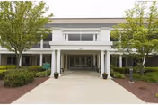 Front exterior view of a building with white columns and a covered entrance, flanked by green trees and shrubs on either side of a wide concrete pathway.