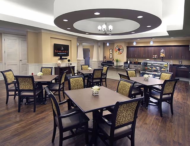 Dining/common room with multiple square tables and patterned chairs, a serving counter and display case beneath a circular recessed ceiling feature.