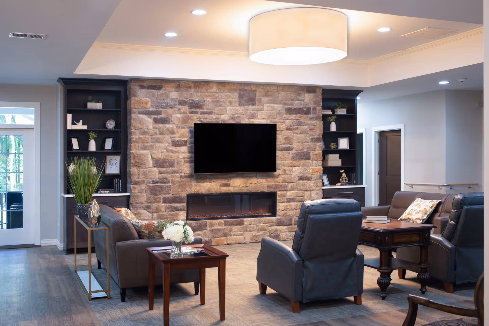Cozy lounge with a stone fireplace and wall-mounted TV flanked by bookshelves, surrounded by leather chairs and side tables.
