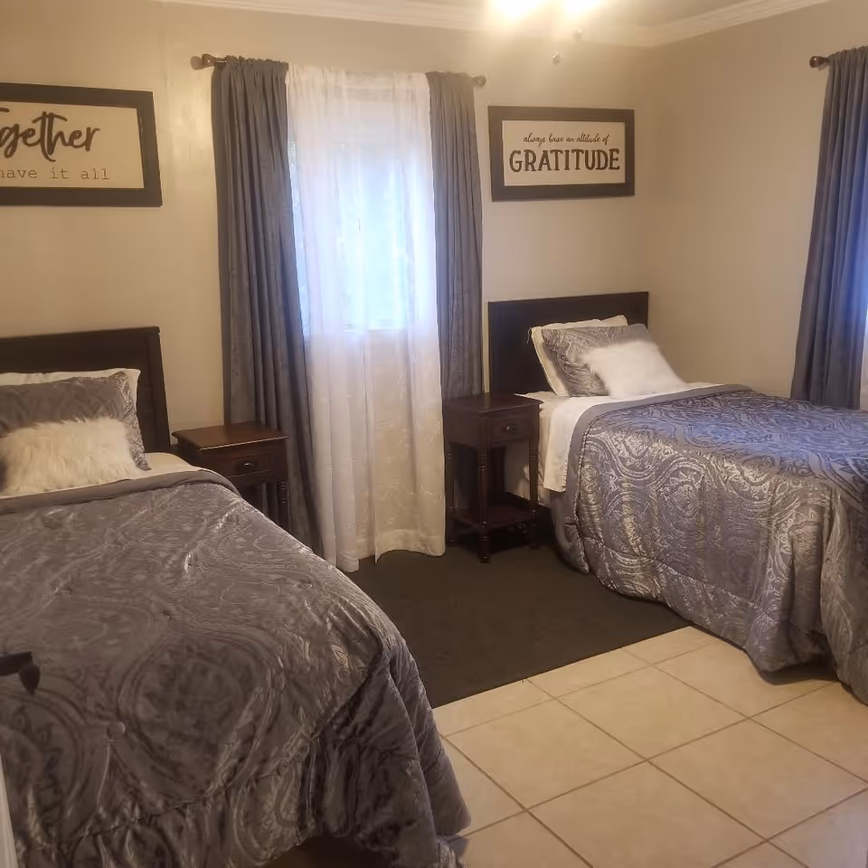 Two twin beds with gray bedspreads and nightstands in a neatly decorated bedroom with curtains and wall art.