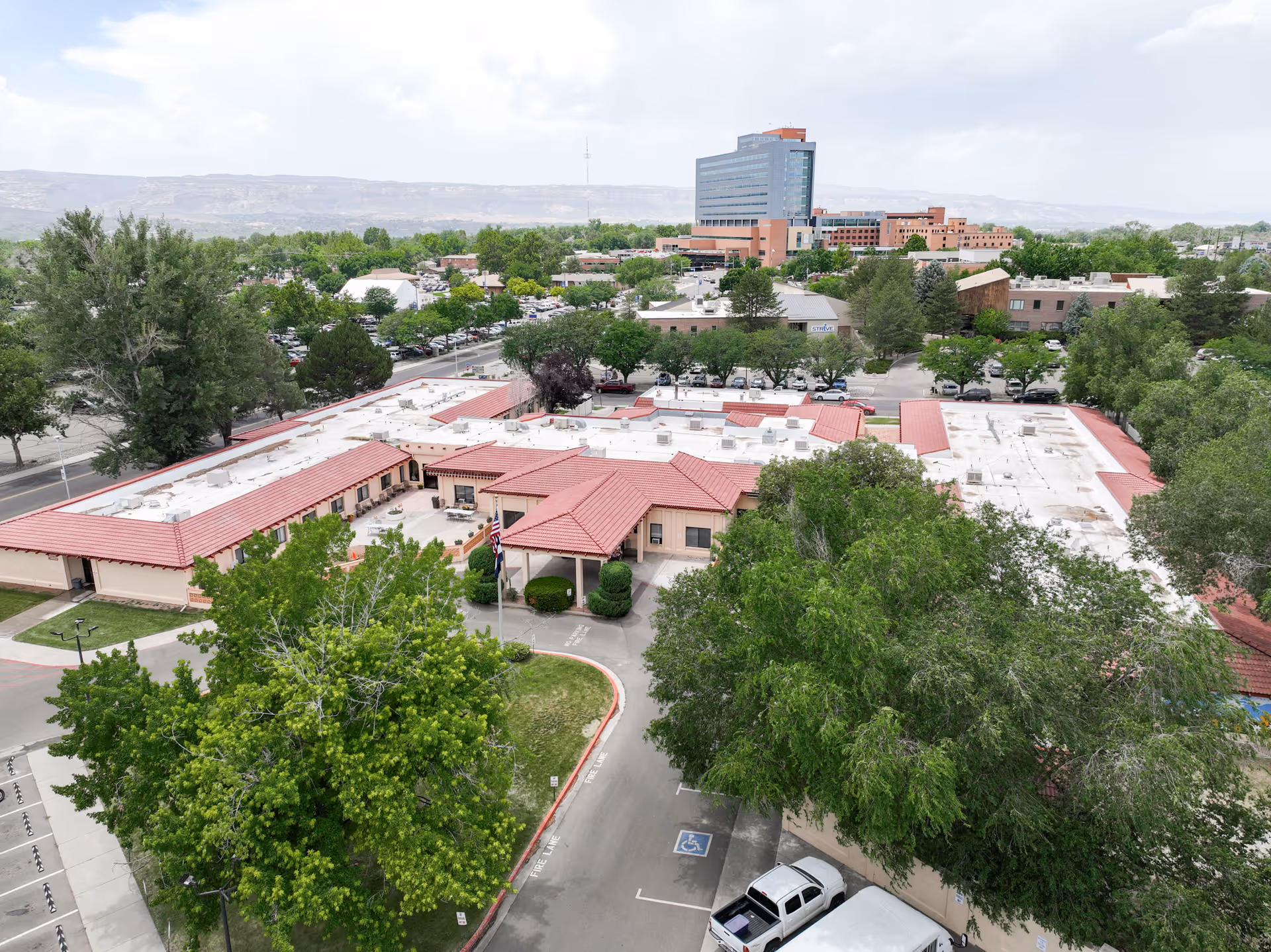 Aerial view of La Villa Grande Care Center showing a single-story building with red-tiled roofs surrounded by trees and parking areas. In the background, there are other buildings and a large multi-story building with mountains visible in the distance under a cloudy sky.