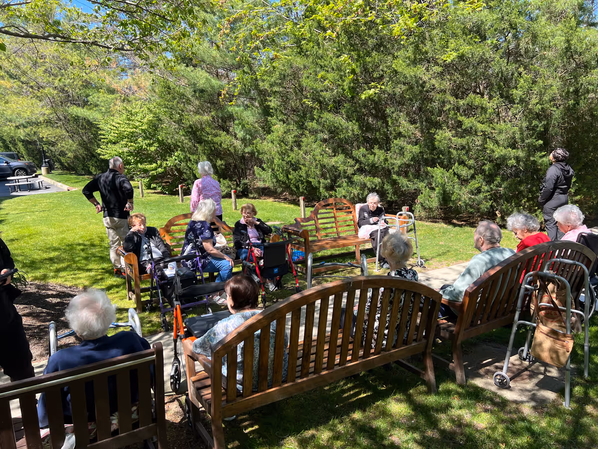 A group of elderly people sitting on wooden benches and chairs in a sunny outdoor garden area surrounded by green trees and grass. Some individuals use walkers and are engaged in conversation or relaxing. Two people are standing nearby.