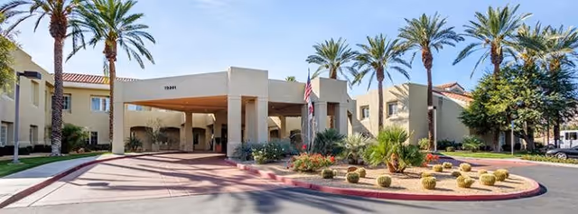Exterior view of Bayshire Rancho Mirage facility entrance with a covered drop-off area, surrounded by palm trees, desert landscaping with cacti, and a clear blue sky.