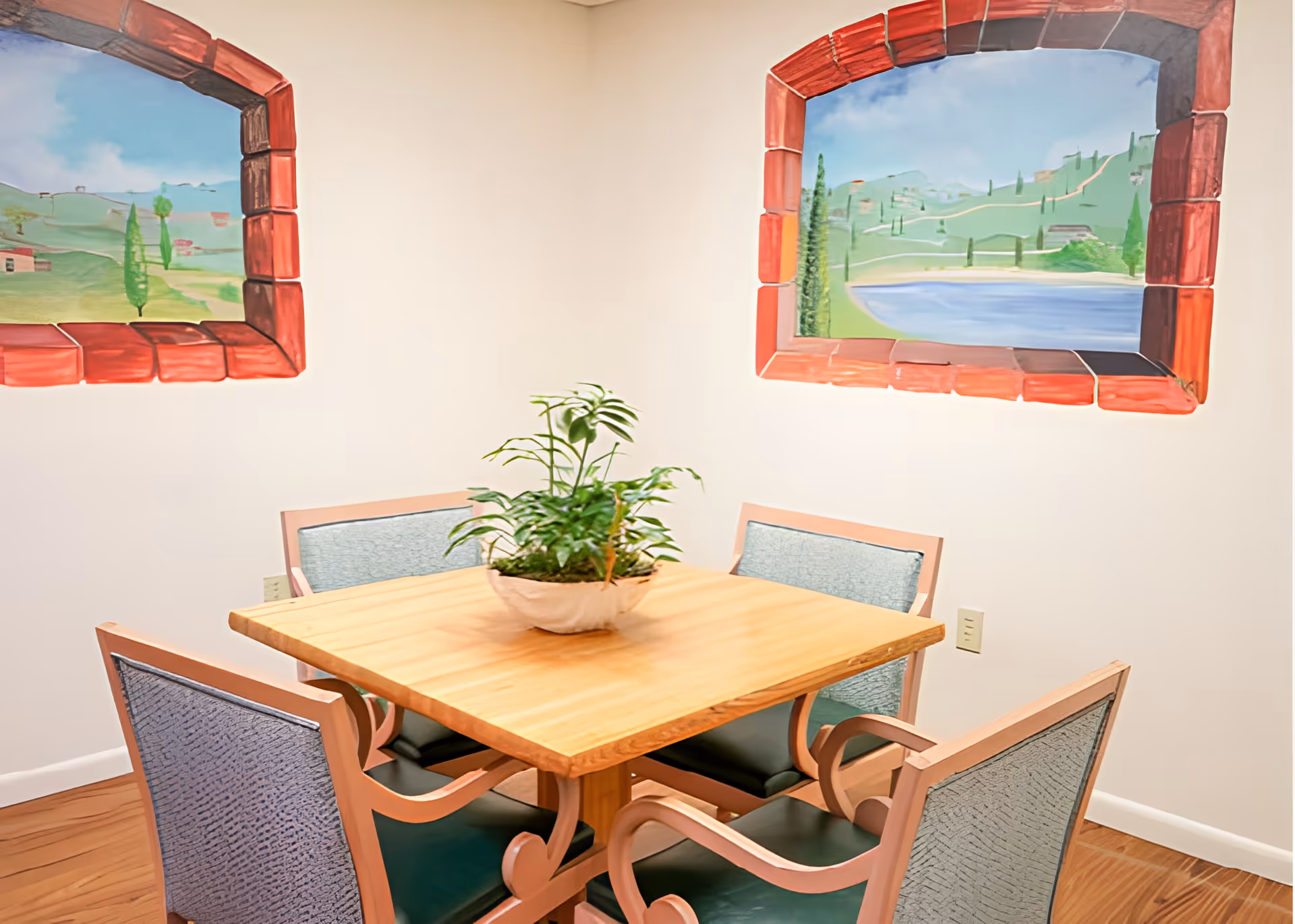 A small square wooden table with a potted green plant in the center, surrounded by four cushioned chairs with light blue upholstery. The walls are white and feature two painted murals designed to look like windows with red brick frames, showing scenic landscapes with greenery and water.