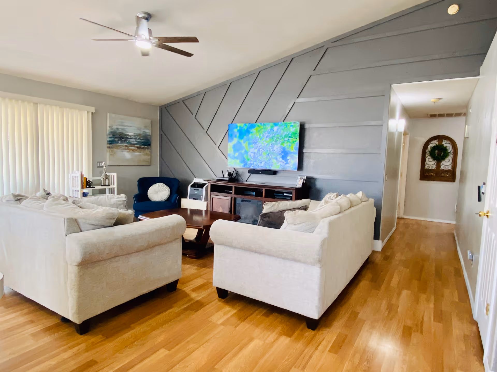 A bright living room with light wood flooring, two beige sofas facing a dark wood coffee table, and a navy blue armchair with a white round pillow. A flat-screen TV is mounted on a gray accent wall with geometric patterns. A ceiling fan with lights is installed on the white ceiling. There is a hallway visible to the right with a decorative wall hanging and a wreath.
