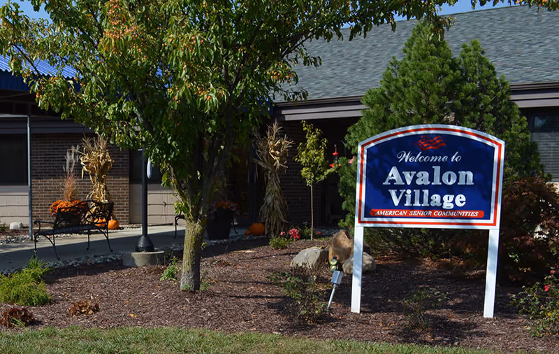 Outdoor view of Avalon Village senior living facility entrance with a blue and white welcome sign, trees, plants, and a bench decorated with autumn-themed items including pumpkins and dried corn stalks.