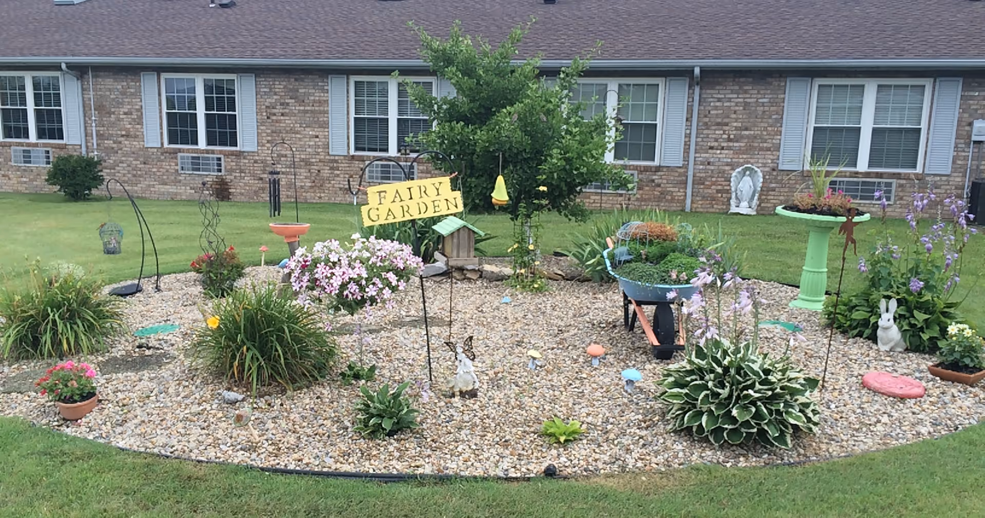 A small decorative garden area labeled 'Fairy Garden' with various plants, flowers, garden ornaments, and a small birdhouse. The garden is surrounded by grass and is in front of a brick building with multiple windows.