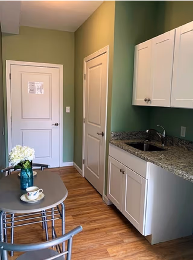 Small kitchen area with green walls, white cabinets, a granite countertop with a sink, and a small table with two metal chairs. On the table, there is a blue vase with white flowers and a teacup with a saucer. Two closed white doors are visible in the background.