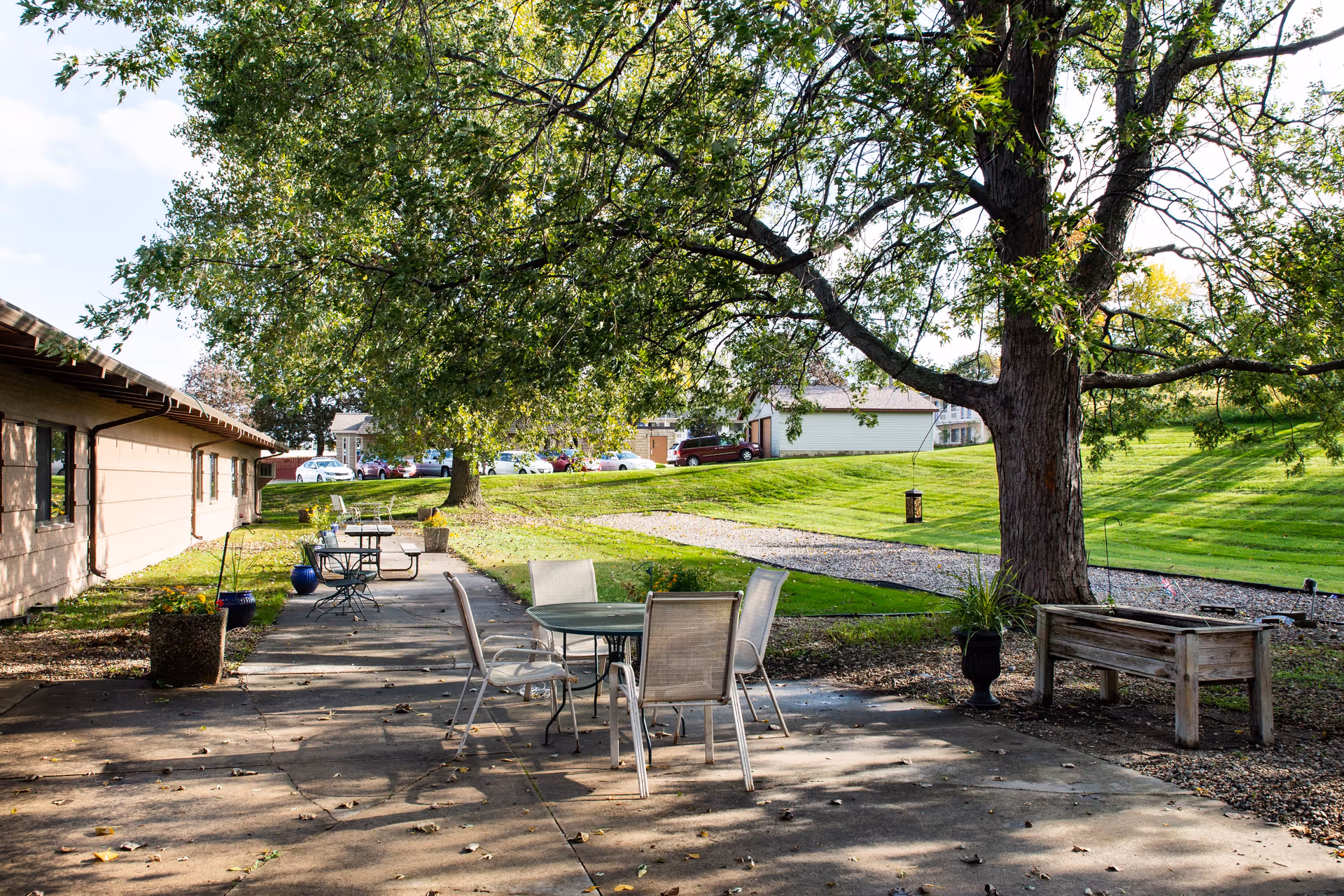 Outdoor patio area with several tables and chairs under large trees providing shade. The patio is adjacent to a building with windows, and there is a grassy area with a gravel path and parked cars in the background.