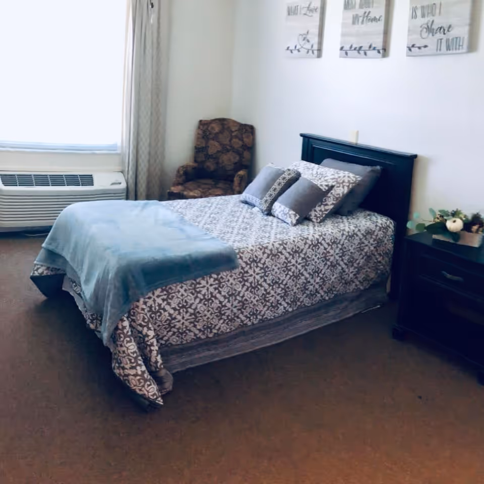 A neatly made bed with patterned bedding and multiple pillows in a bedroom. There is a blue throw blanket at the foot of the bed. Next to the bed is a dark wooden nightstand with a small decorative plant. In the corner near the window, there is a patterned armchair. The window has light-colored curtains and a heating/cooling unit below it. Three framed wall decorations with text hang above the bed.