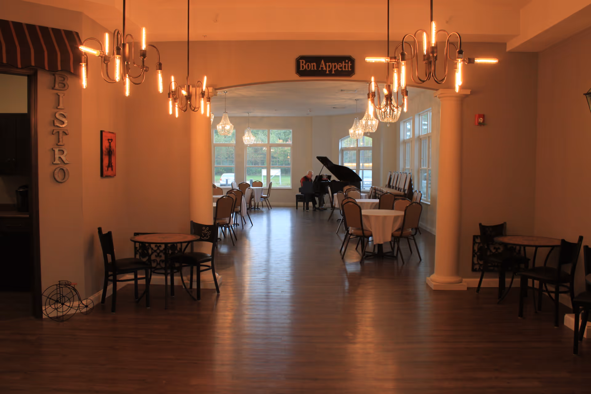 Dining room interior with round tables and chairs, hanging chandeliers, and a grand piano beneath a 'Bon Appetit' sign.