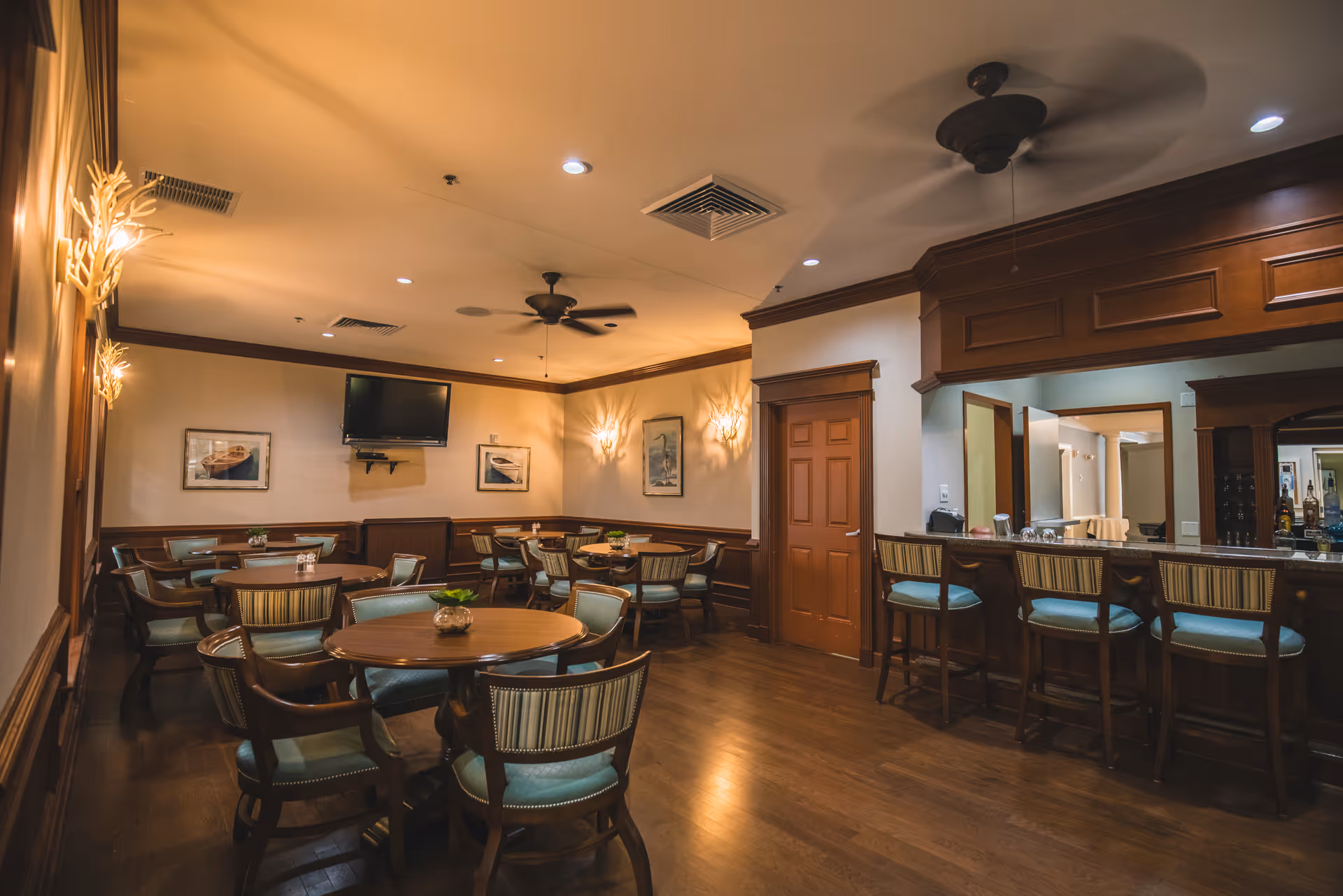 A cozy dining area with several round wooden tables surrounded by cushioned chairs. The room features warm lighting, wooden paneling, a ceiling fan, and a bar counter with high chairs. There is a wall-mounted television and framed pictures on the walls.
