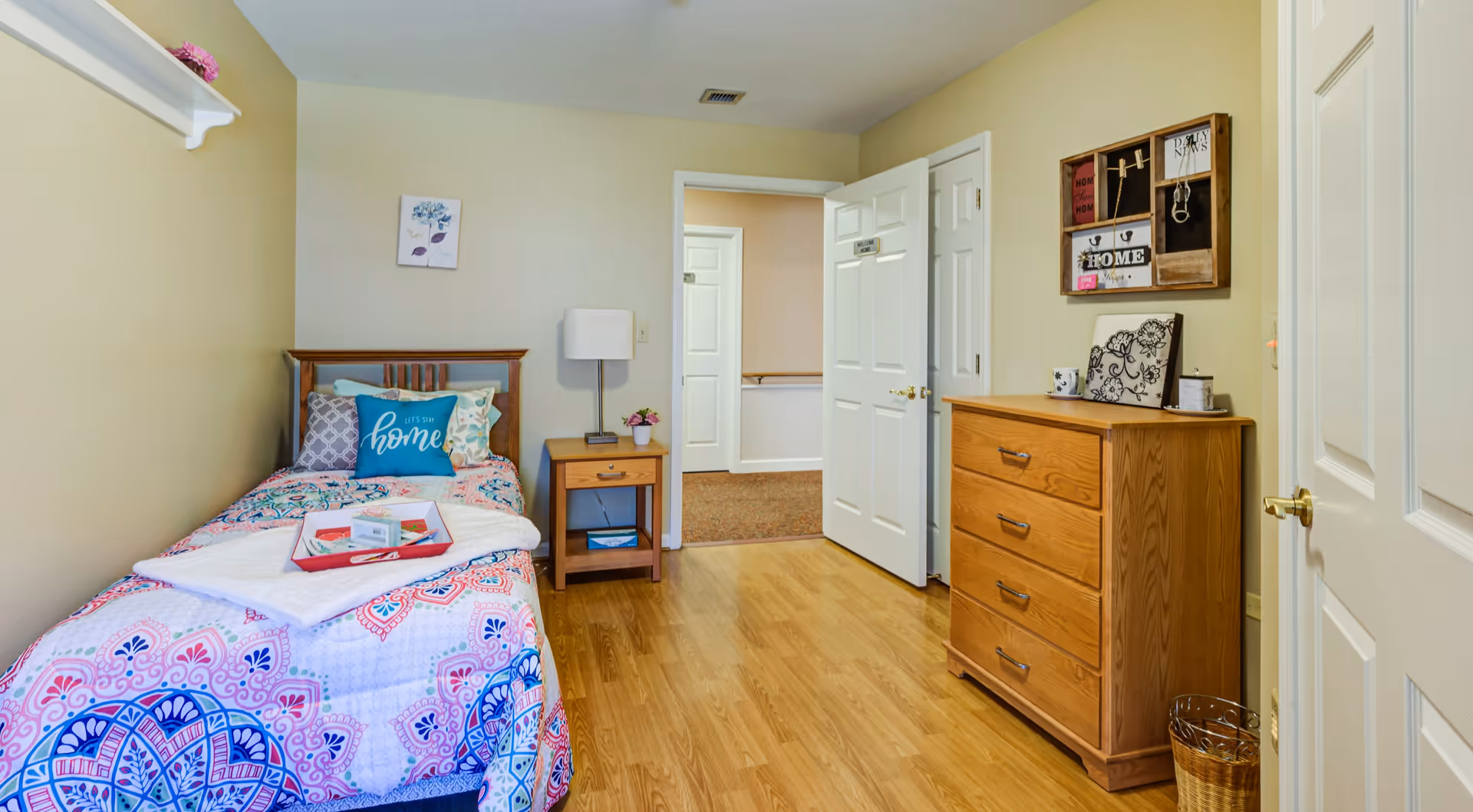 A small, cozy bedroom with a single bed covered in a colorful patterned quilt and multiple pillows, including one with the word 'home'. Next to the bed is a wooden nightstand with a lamp and a small plant. Across from the bed is a wooden dresser with decorative items on top and a wall-mounted organizer above it. The room has light-colored walls, wooden flooring, and an open door leading to a hallway.