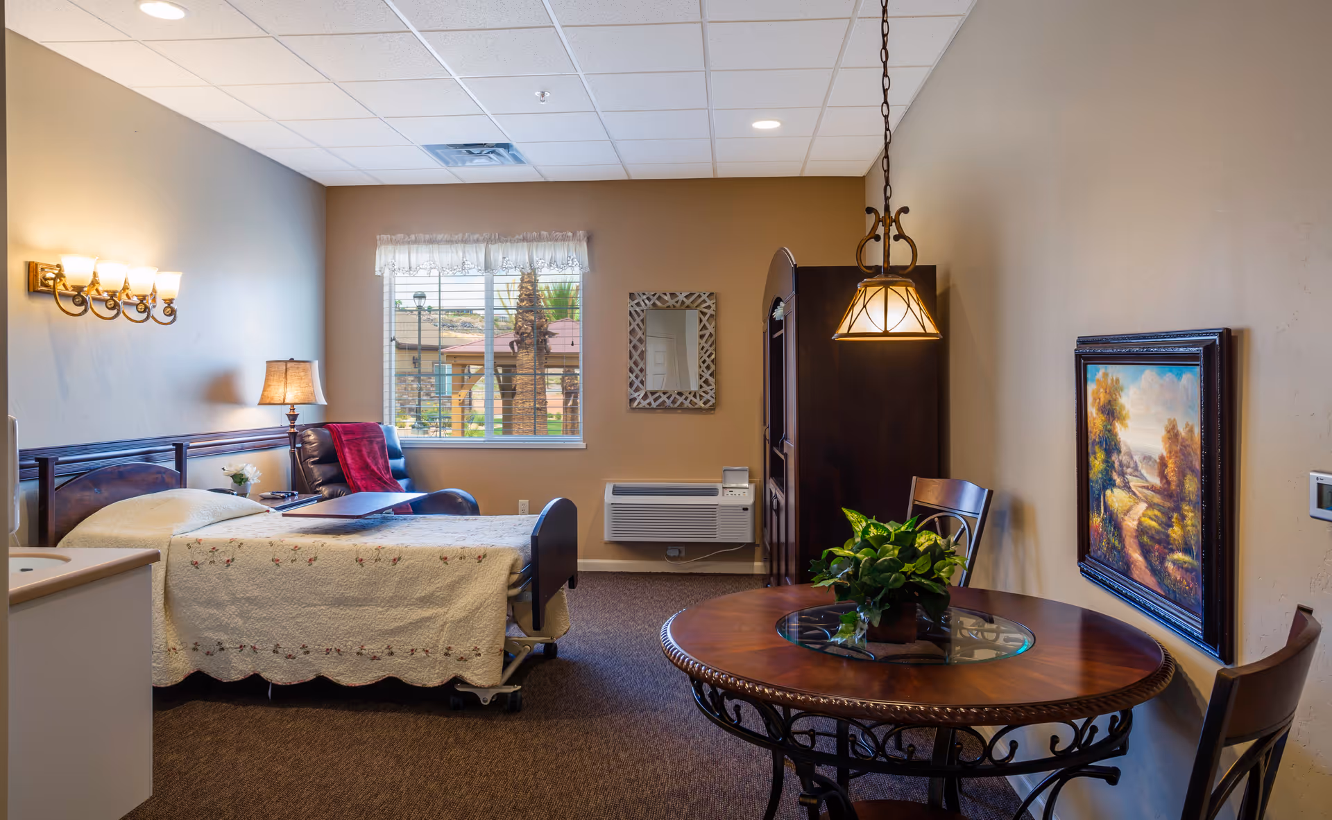 Private senior living room with a bed and bedside lamp, a recliner by a window, a round wooden table with chairs, and wall art.
