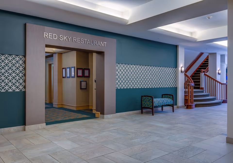 Interior view of a senior living facility hallway featuring an entrance to the Red Sky Restaurant with a decorative archway. The walls are painted teal with a patterned horizontal stripe. There is a small upholstered bench with a floral pattern near a curved wooden staircase with carpeted steps. The floor is tiled in light stone.