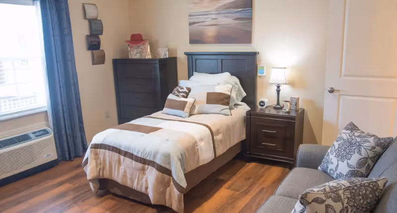 A cozy bedroom with a single bed covered in a beige and brown striped comforter and multiple pillows. Next to the bed is a dark wooden nightstand with a lamp, clock, and small decorative items. A matching dark wooden dresser with a red hat and other items on top is against the wall. There is a window with blue curtains and an air conditioning unit below it. A gray couch with floral patterned pillows is partially visible on the right side. The room has wooden flooring and a door on the right wall.
