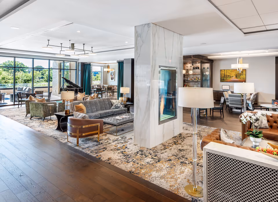 A spacious and elegantly furnished living area in a senior living facility featuring a modern fireplace with marble surround in the center, multiple seating arrangements including sofas and armchairs, a grand piano near large windows with a view of greenery outside, and a dining area with tables and chairs in the background.