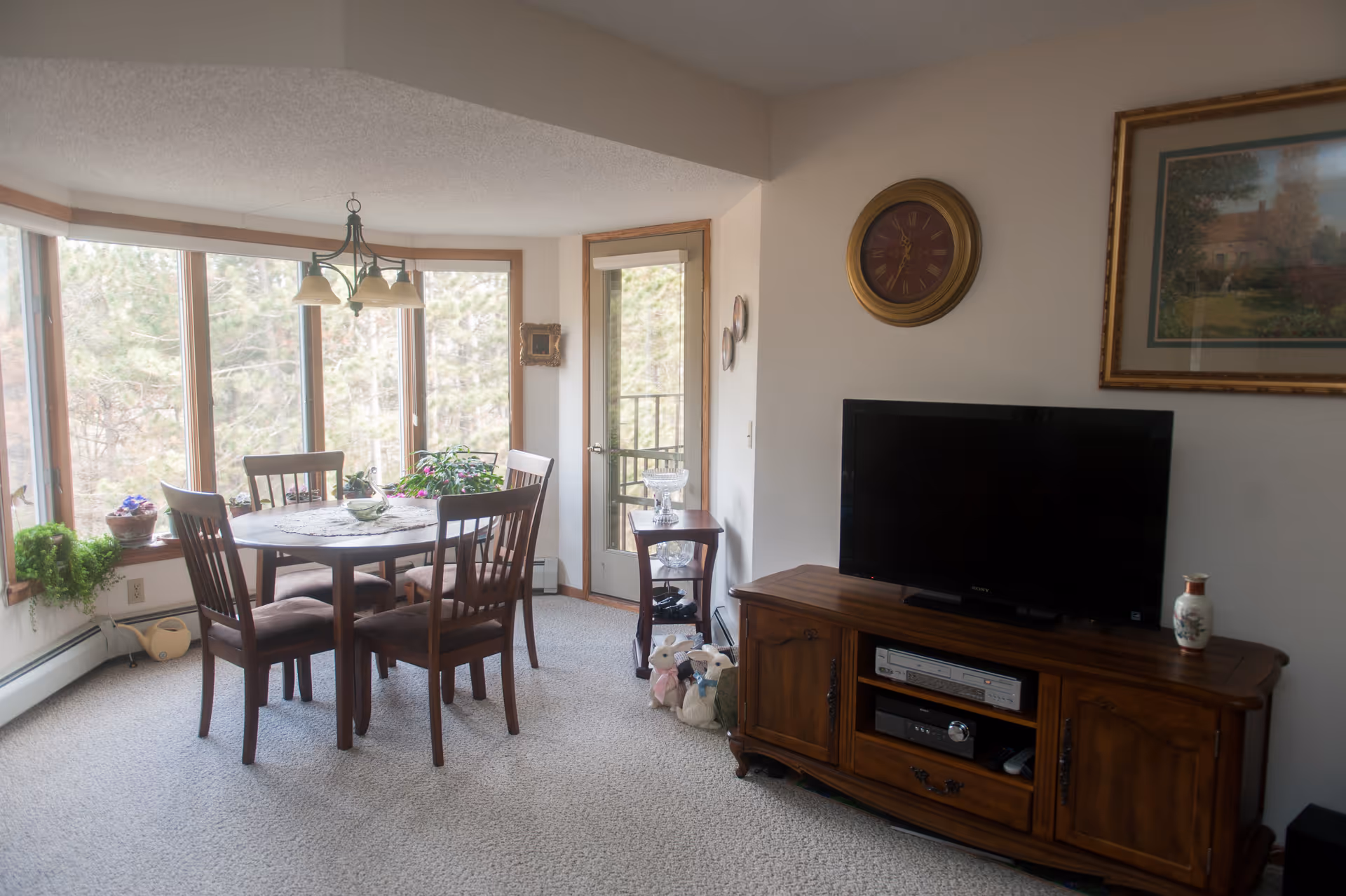 A cozy living and dining area with a round wooden dining table and four chairs near large windows letting in natural light. A wooden TV stand holds a flat-screen television and decorative items, with a wall clock and framed artwork above it. There are plants on the windowsill and a small side table near a glass door leading outside.