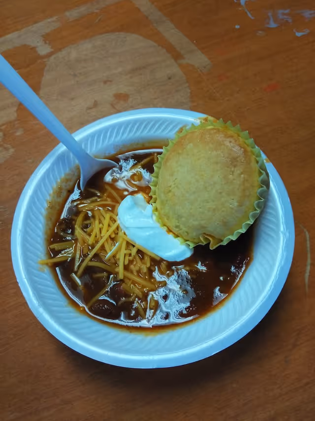 A white disposable bowl containing chili topped with shredded cheddar cheese and a dollop of sour cream, accompanied by a cornbread muffin in a yellow paper liner, with a white plastic spoon resting inside the bowl on a wooden table.