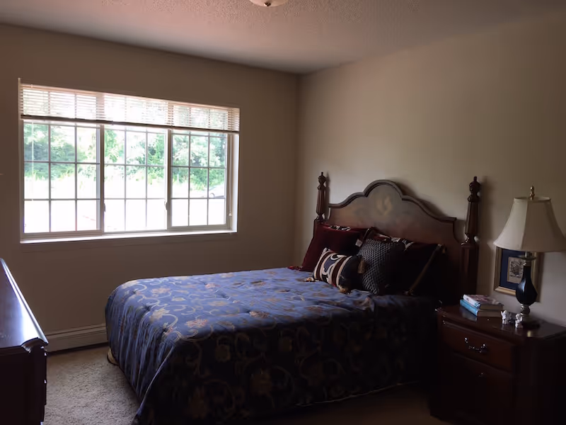 A bedroom with a large window letting in natural light. The room features a wooden bed with an ornate headboard, covered with a dark blue patterned bedspread and several decorative pillows. There is a wooden nightstand with a lamp, books, and small decorative items next to the bed. The walls are plain and light-colored.