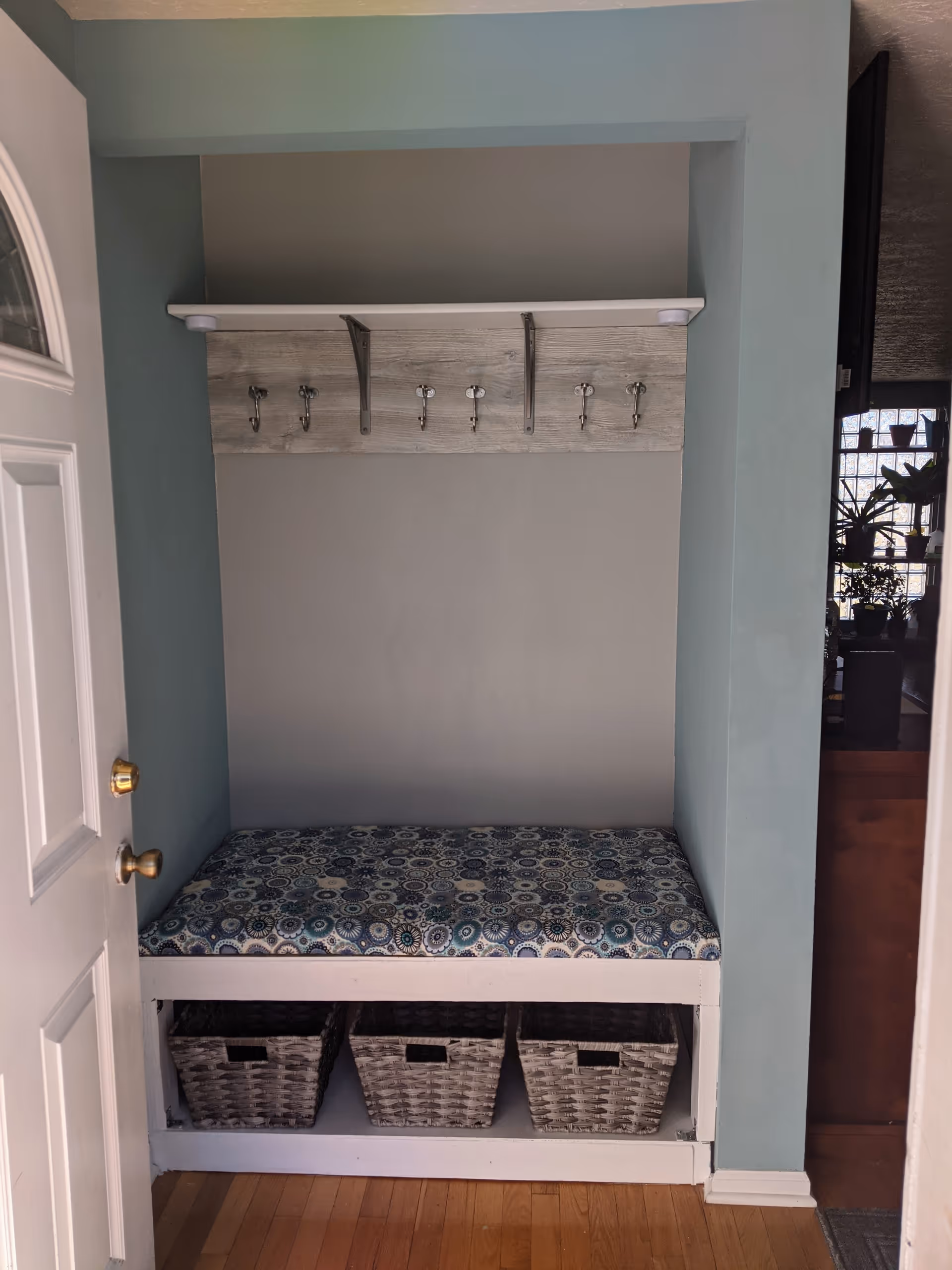 A small entryway nook with a cushioned bench featuring a blue and white patterned fabric. Above the bench is a wooden panel with six metal hooks for hanging items. Below the bench are three woven storage baskets. The walls are painted light blue and the floor is hardwood. A partially open white door with a brass doorknob is visible on the left side.