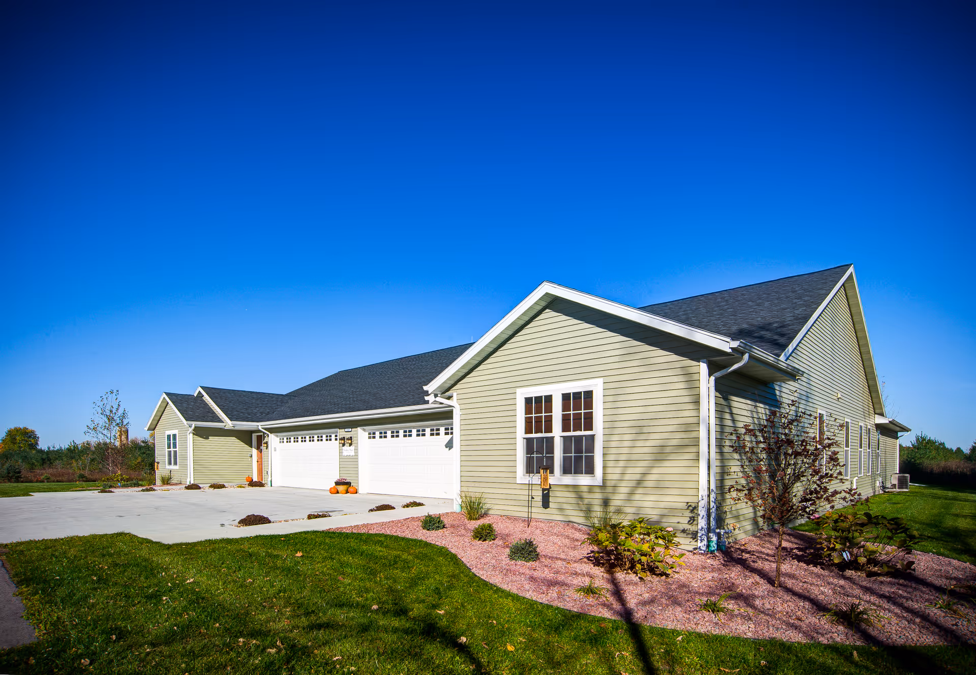 Exterior view of a single-story residential building with light green siding, white trim, and a dark shingled roof under a clear blue sky. The building has multiple garage doors and a landscaped area with small plants and mulch in front.