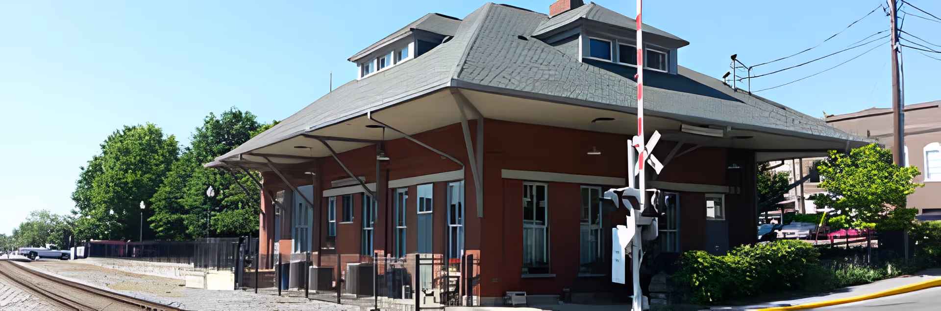 Exterior view of a vintage-style train station building with a sloped roof, multiple windows, and a railroad crossing sign in front. The scene includes train tracks on the left and green trees surrounding the area under a clear blue sky.