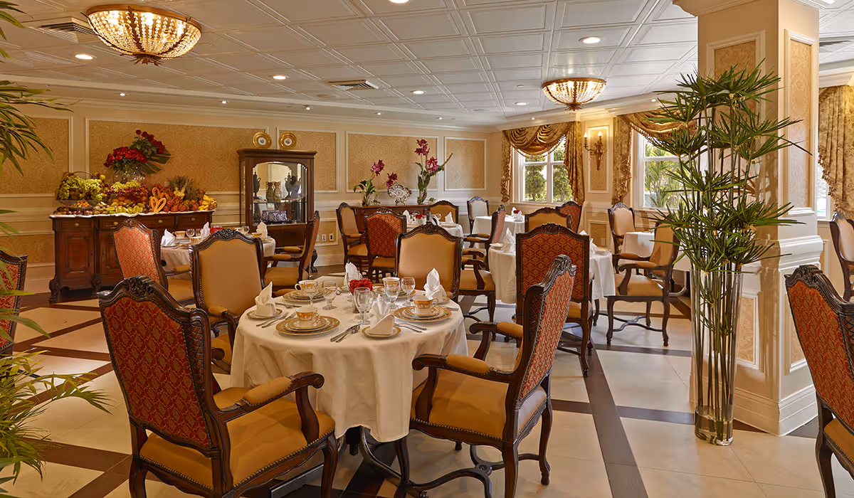 Elegant dining room with round tables covered in white tablecloths, set with fine china, glassware, and napkins. The room features ornate chairs with red and gold upholstery, decorative plants, large windows with draped curtains, and warm lighting from chandeliers and wall sconces.