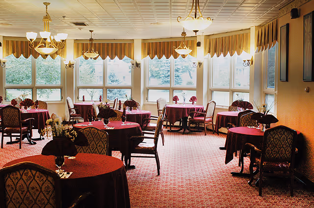 A dining room with multiple round tables covered in burgundy tablecloths, each set with folded napkins and small floral centerpieces. The room has large windows with striped valances allowing natural light to enter, and several chandeliers hanging from the ceiling. The carpet is patterned in red tones, and the chairs have cushioned seats and backs with a decorative design.