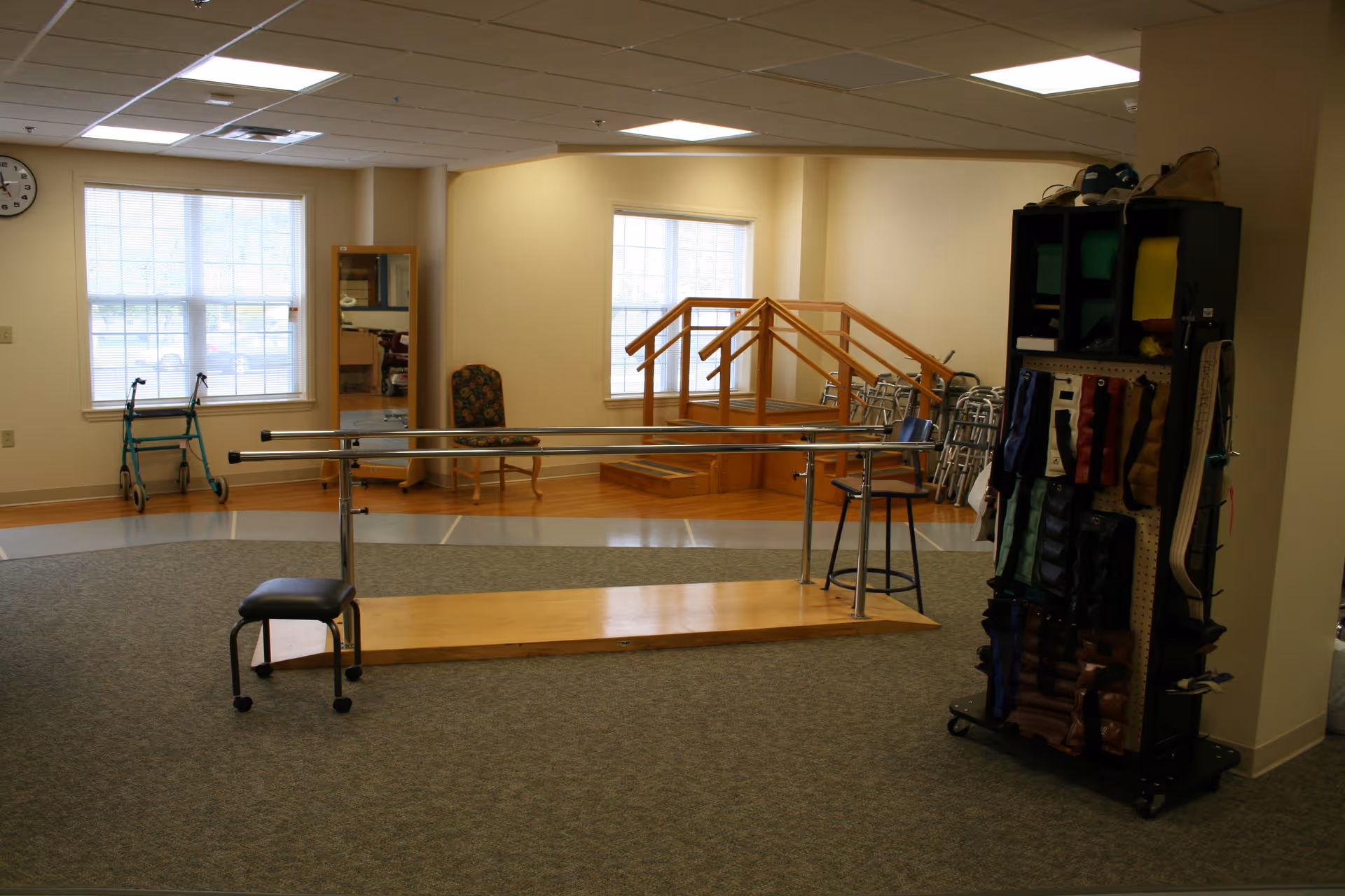A rehabilitation therapy room with parallel bars for walking exercises, a stool, a chair, a mirror, a walker, several wheelchairs, and a rack holding various therapy equipment and braces. The room has large windows letting in natural light and a clock on the wall.